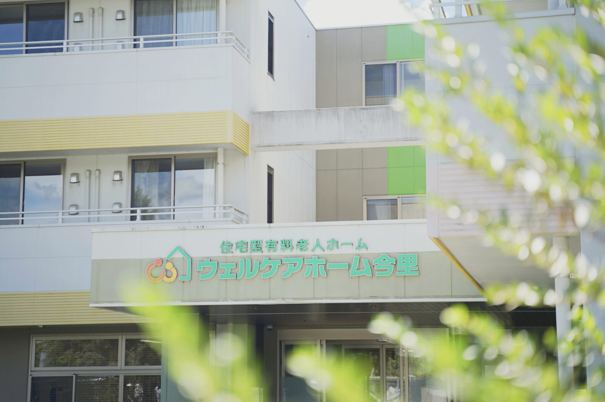 介護施設の建物外観。白と黄色を基調とした多層階の建物で、緑のアクセントが見られます。